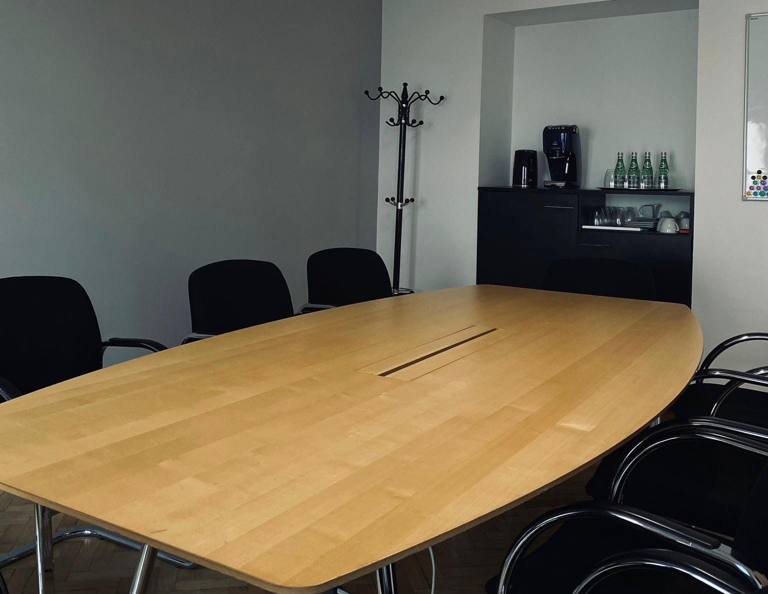 Jasna sala konferencyjna z dużym, drewnianym stołem, prawdopodobnie Vitra Medamorph. Wokół stołu czarne krzesła. W tle szafka z ekspresem do kawy i napojami.