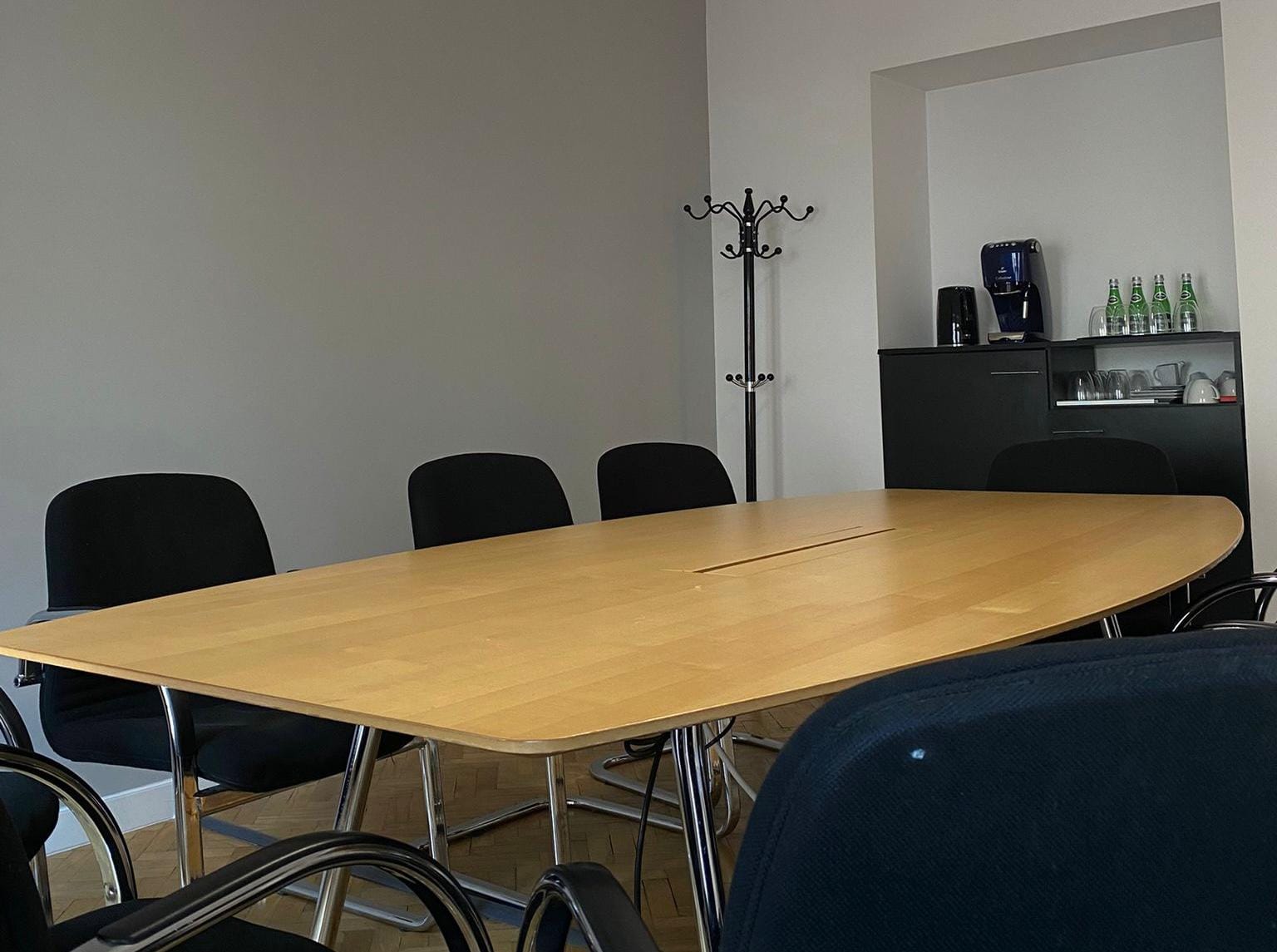 Sala konferencyjna: duży drewniany stół, czarne krzesła, wieszak na ubrania. W tle szafka z ekspresem i szklankami.