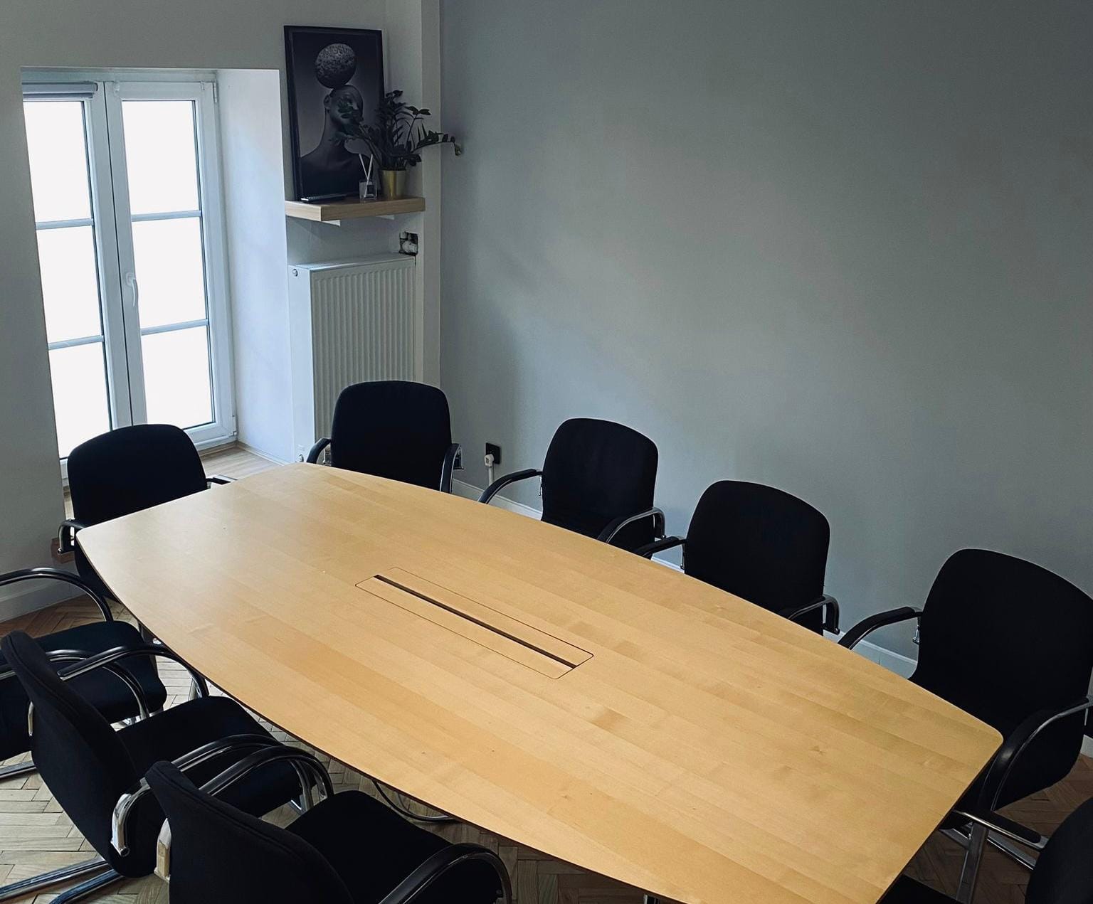 Sala konferencyjna z jasnym drewnianym stołem i czarnymi krzesłami. W tle okno i półka z ozdobami. Szare ściany.