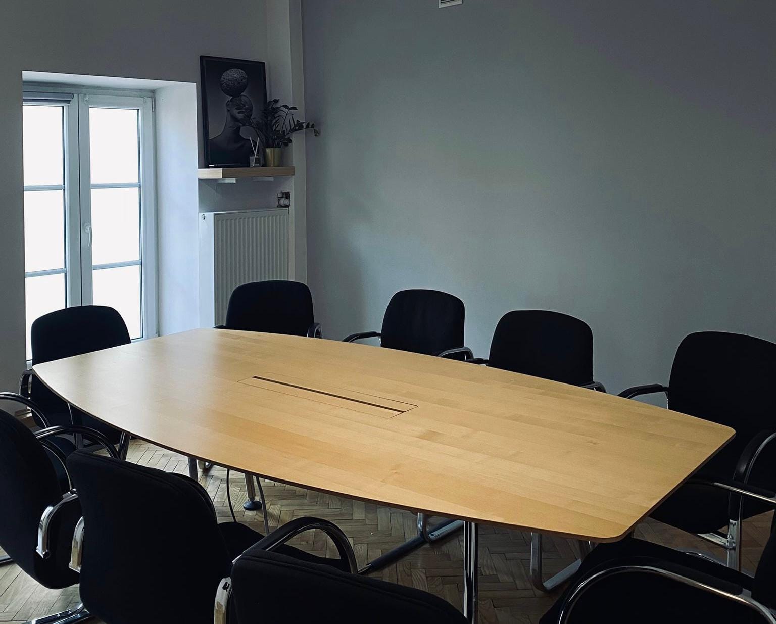 Jasna sala konferencyjna. Duży, owalny, drewniany stół otoczony czarnymi krzesłami. W tle okno i kaloryfer.