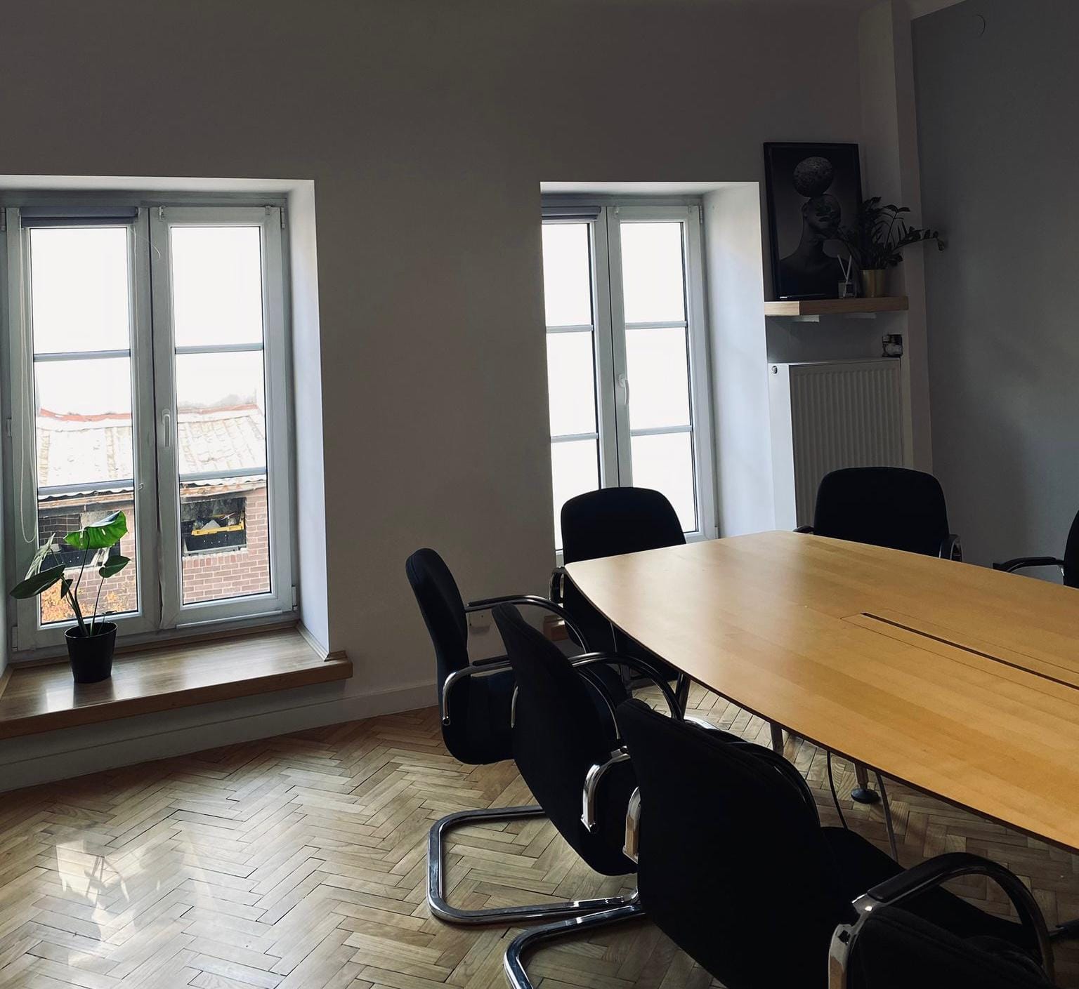 Sala konferencyjna z dużym drewnianym stołem, czarnymi krzesłami i dwoma oknami. Na parapecie roślina doniczkowa.