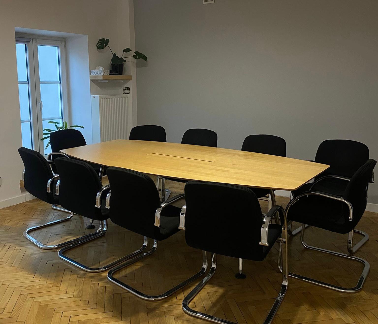 : Sala konferencyjna z dużym, jasnym, drewnianym stołem i czarnymi krzesłami na chromowanych nogach. W tle okno i roślina.