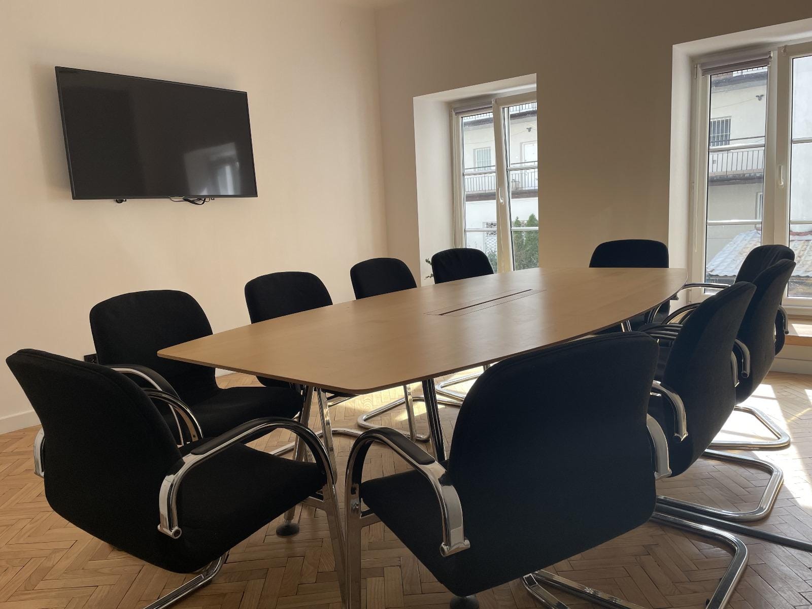 Sala konferencyjna: prostokątny, drewniany stół, czarne krzesła. W tle wiszący na ścianie telewizor i okno.